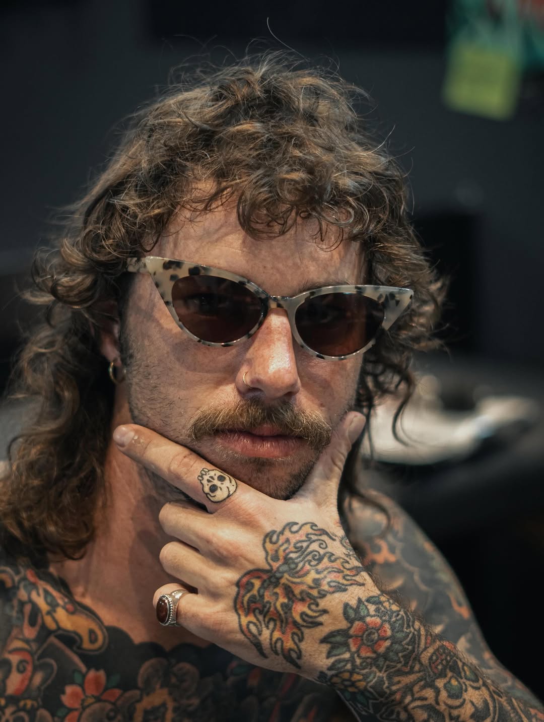Man with long curly hair wearing sunglasses displays extensive colorful floral and decorative tattoos covering his arms and chest.