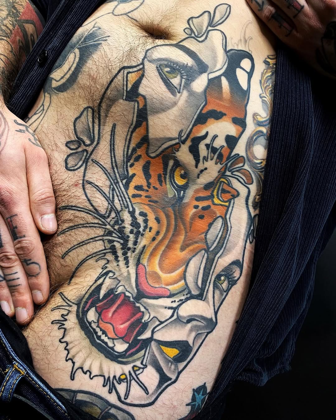 Colorful tiger head tattoo on the ribs with black outlines, orange and yellow flames, red mouth, and decorative floral elements surrounding the design.