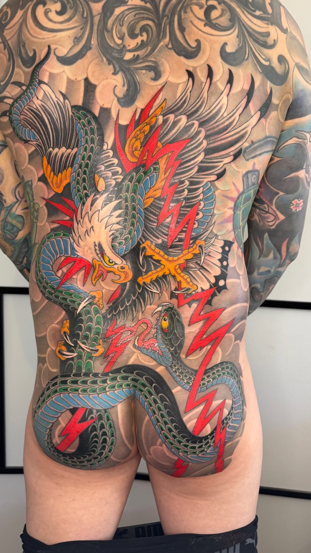 Back piece tattoo featuring a winged eagle fighting a coiled dragon with red lightning bolts, ornamental scrollwork, green scales, and yellow/orange accents.