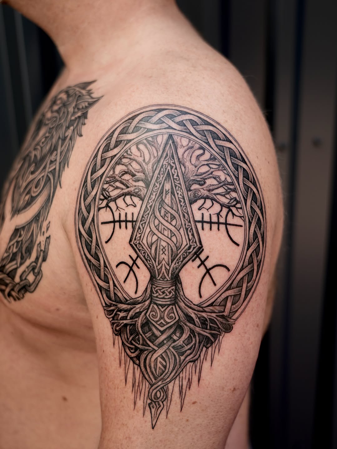 Intricate tree-of-life design with Celtic knotwork border, bare branches, and decorative roots tattooed on upper arm in black ink.