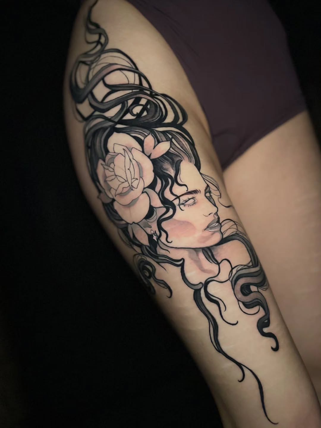 Woman's thigh tattoo featuring a profile of a female face with wavy hair, floral elements, and serpentine design details in black ink with skin-tone shading.