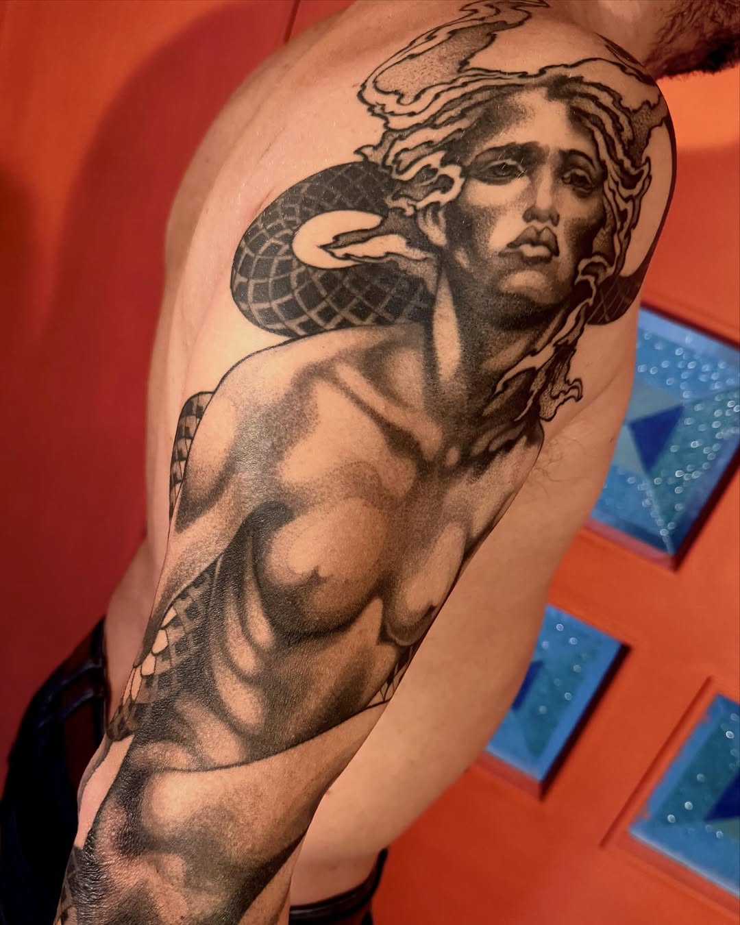 Detailed monochrome shoulder tattoo depicting a classical male figure with winged elements and draped fabric, showcasing shading and anatomical detail.