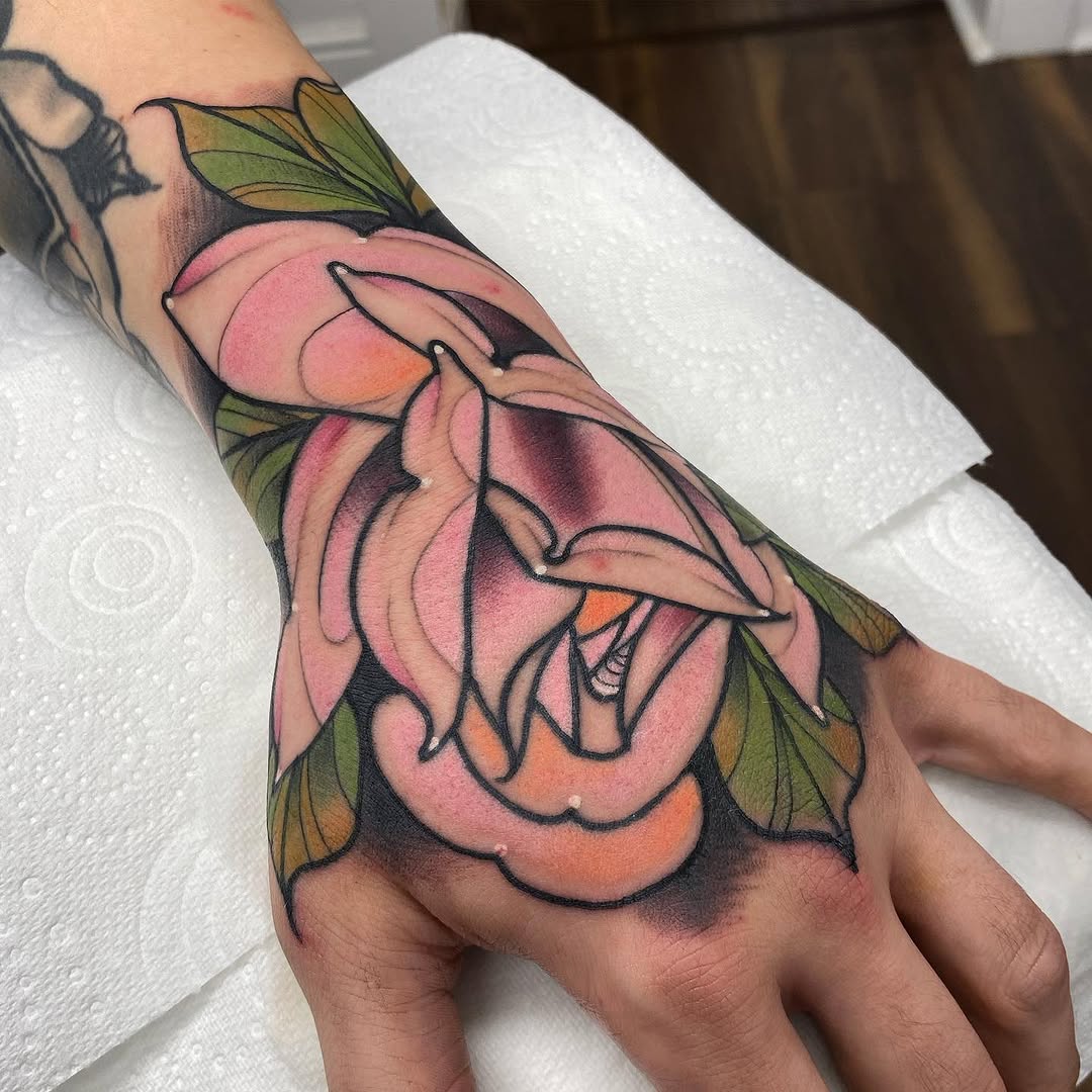 Colorful multi-layered pink and orange flower with green leaves tattooed on the back of a hand, featuring bold black outlines.