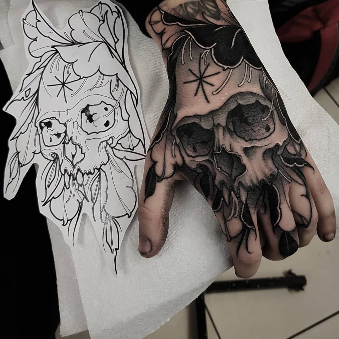 Skull with floral elements tattooed on hand, shown next to inked design sketch on white paper.