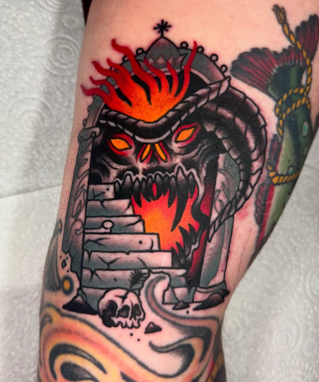Colorful skull tattoo with red and orange flames erupting from the crown, stone structure body, and decorative elements on the arm.