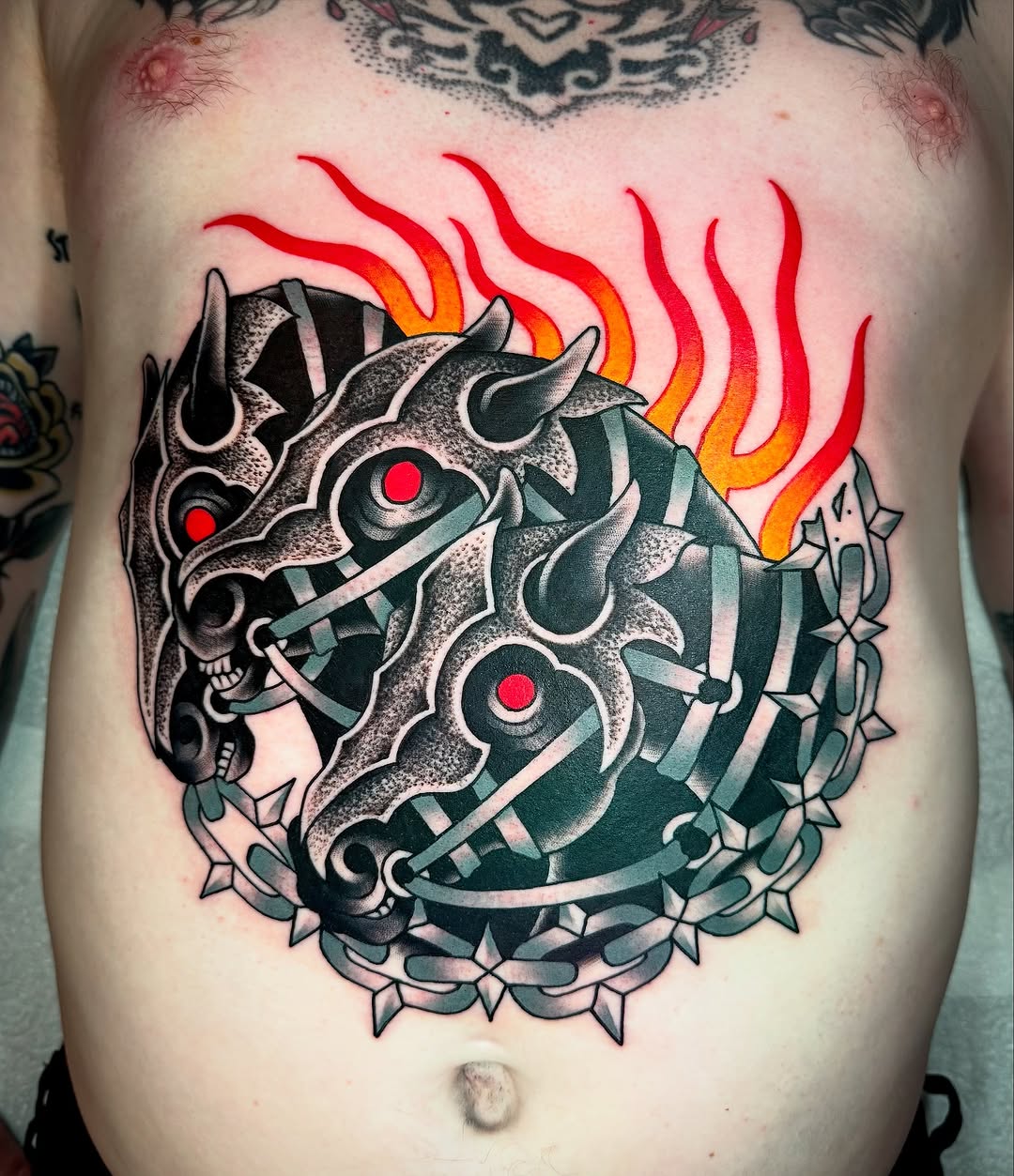 Colorful chest tattoo featuring two demon masks with red eyes, flames above, ornamental borders, and green striping details on bare skin.