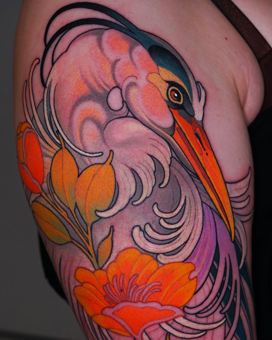 Colorful bird tattoo on upper arm featuring pink, orange, and red plumage with detailed feather work and orange flowers below.