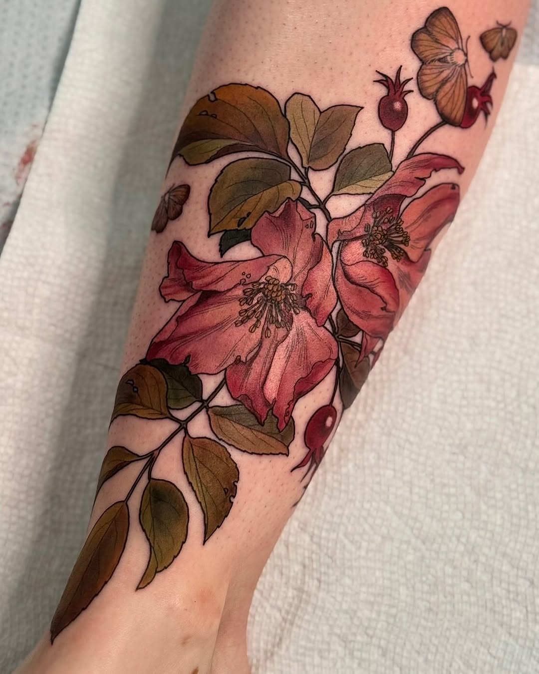 Botanical tattoo on forearm featuring pink flowers with detailed stamens, brown leaves, berries, and butterflies in warm earth tones.