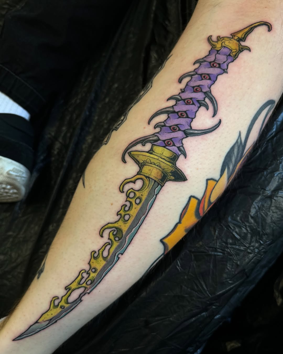 Ornate fantasy dagger tattoo with purple segmented blade, gold decorative handle, and yellow flame accents on the forearm.