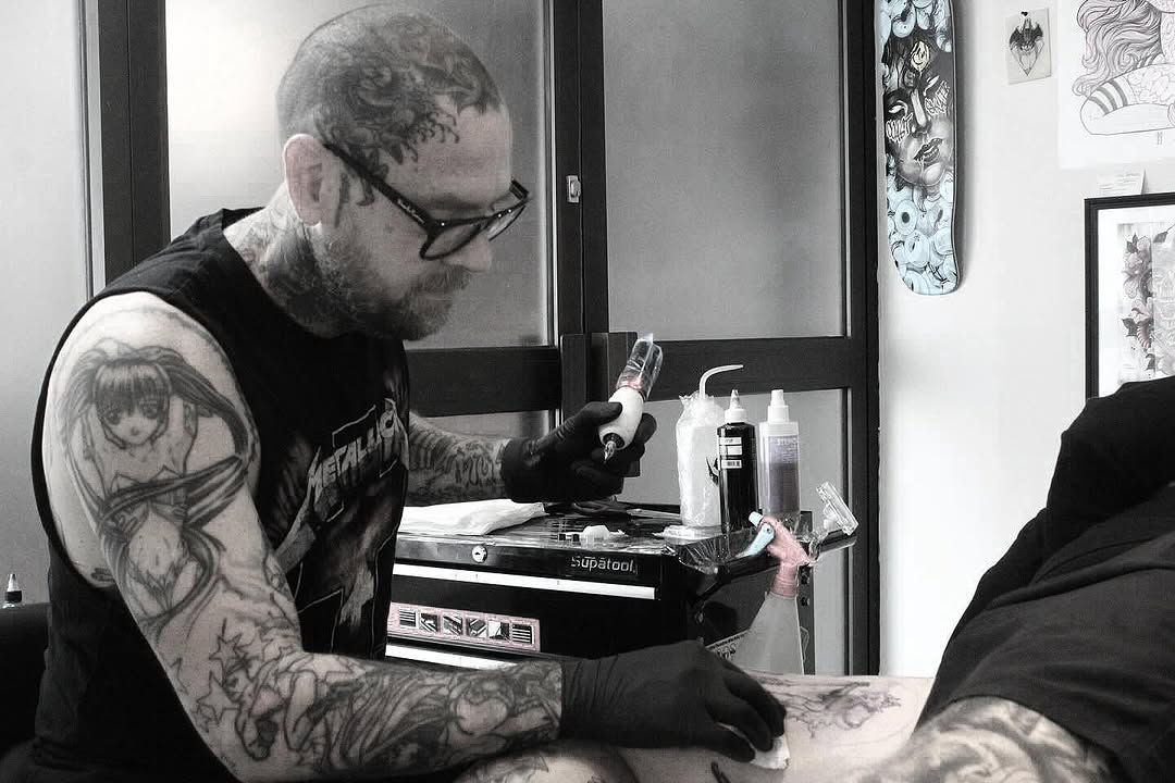 Tattoo artist wearing glasses and black gloves works on a client's arm with a tattoo machine in a studio setting.