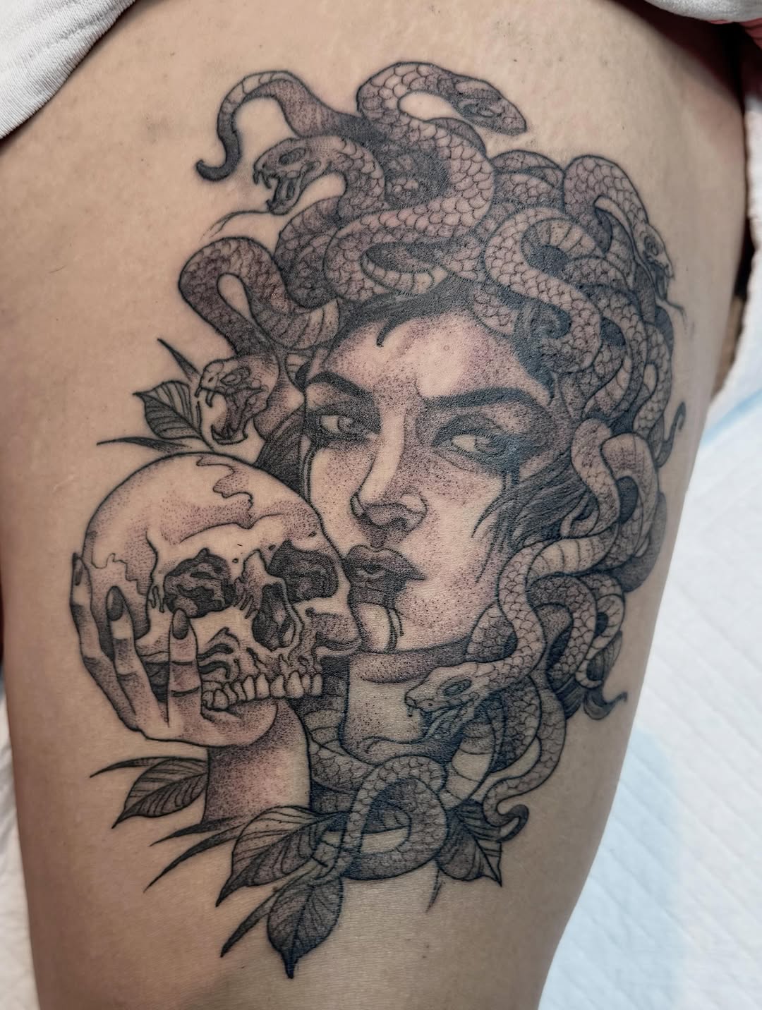 Medusa-inspired portrait tattoo on thigh featuring a woman's face with serpents for hair, a skull held near her face, and botanical elements, rendered in black and grey ink.