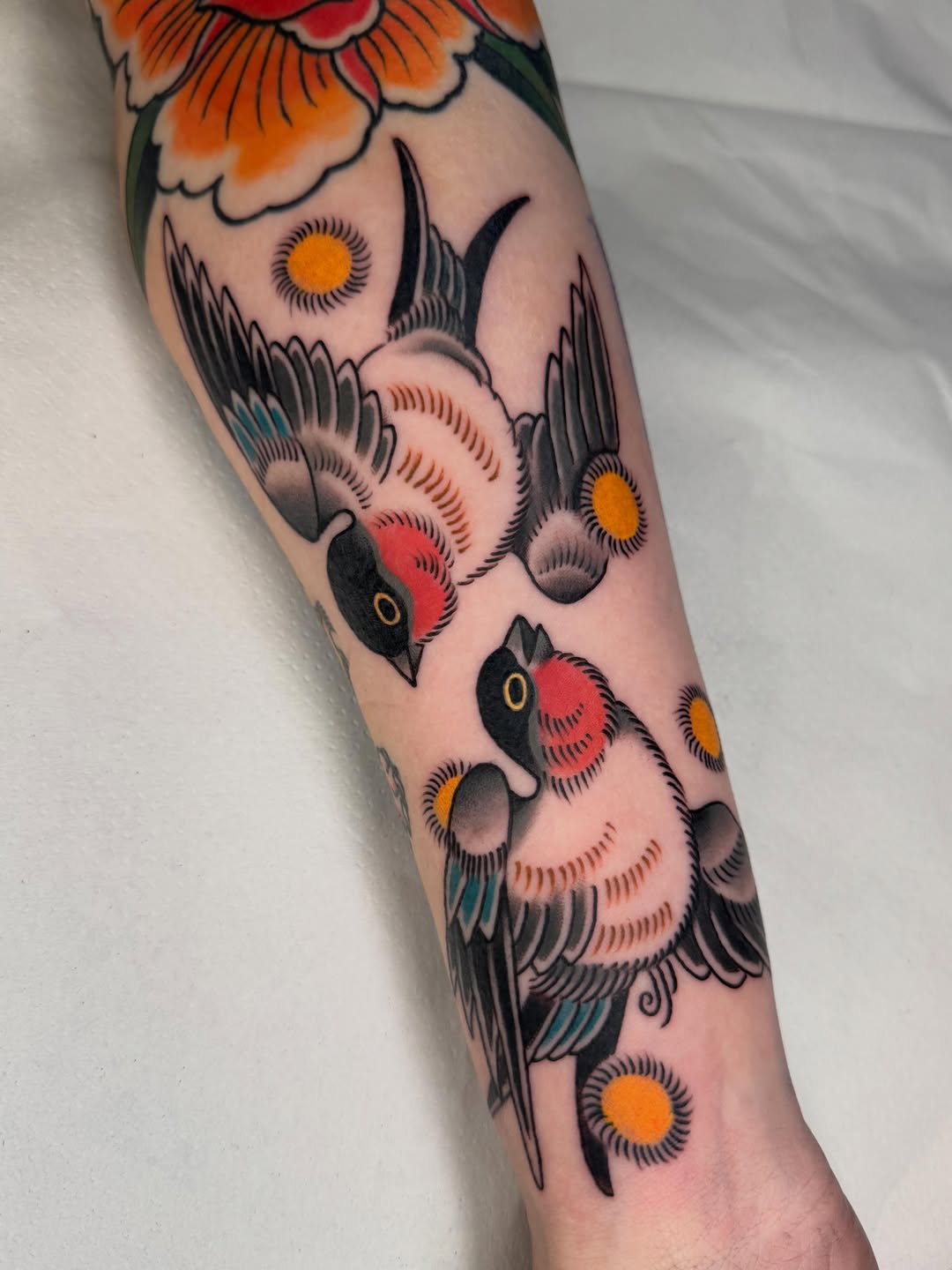 Colorful bird and floral tattoo on forearm featuring multiple owls with orange and red accents, yellow eyes, and a large flower at the top.
