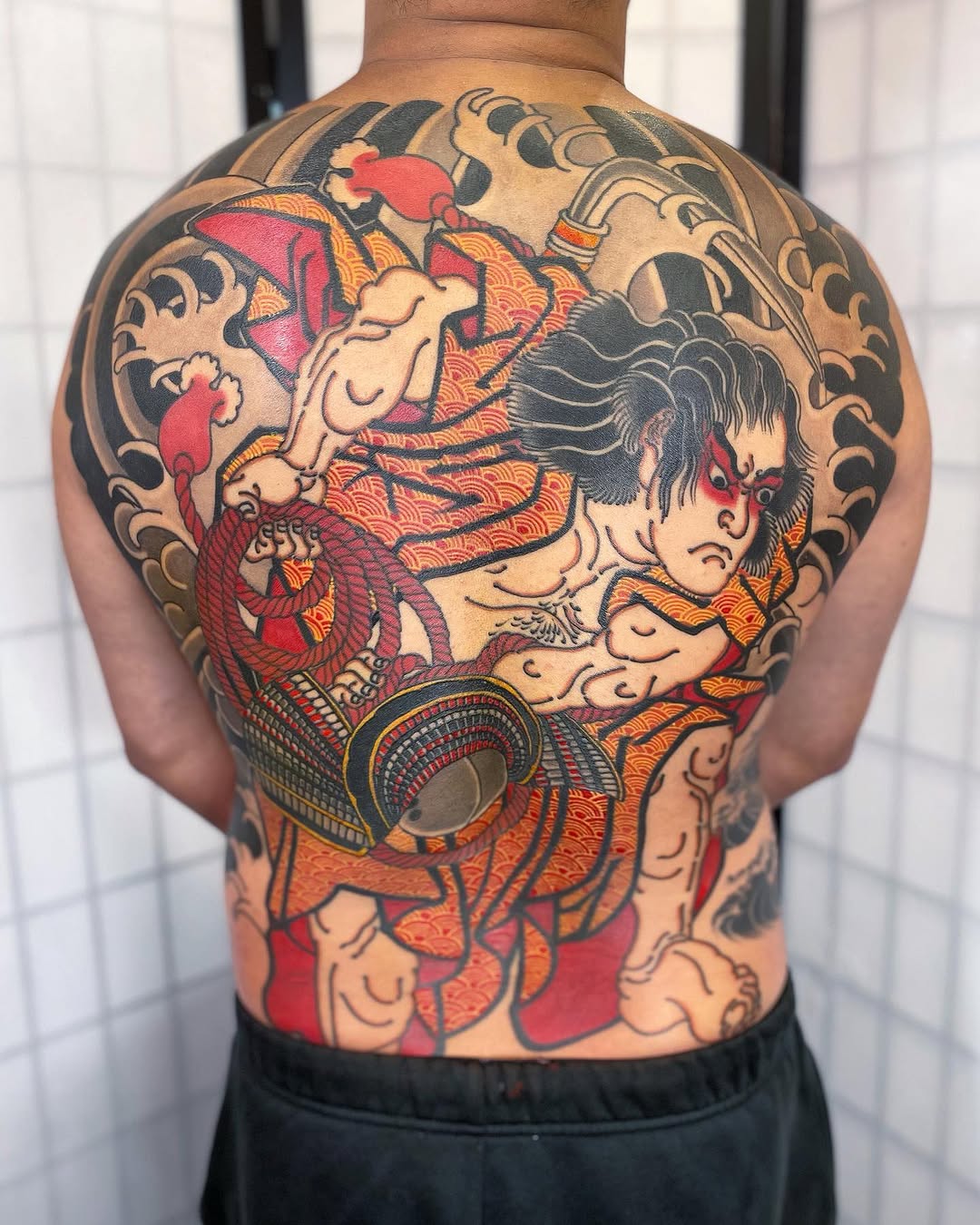 Colorful back tattoo featuring mythological figures, demons, and ornate patterns in red, orange, black, and cream across the upper and lower back.