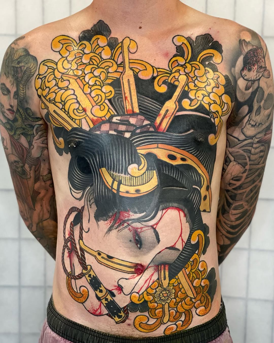 Colorful chest tattoo featuring a stylized face with ornate golden decorative elements, floral motifs, and complementary arm pieces on a shirtless male torso.