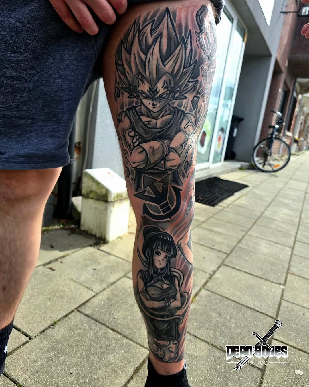 Full leg sleeve tattoo featuring an anime character with spiky hair at top, a female character lower on the calf, rendered in black ink on tan skin.
