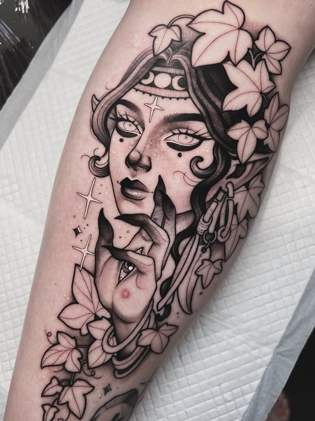 Two overlapping female faces with closed eyes, floral elements, crosses, and decorative details tattooed on a forearm in black ink.