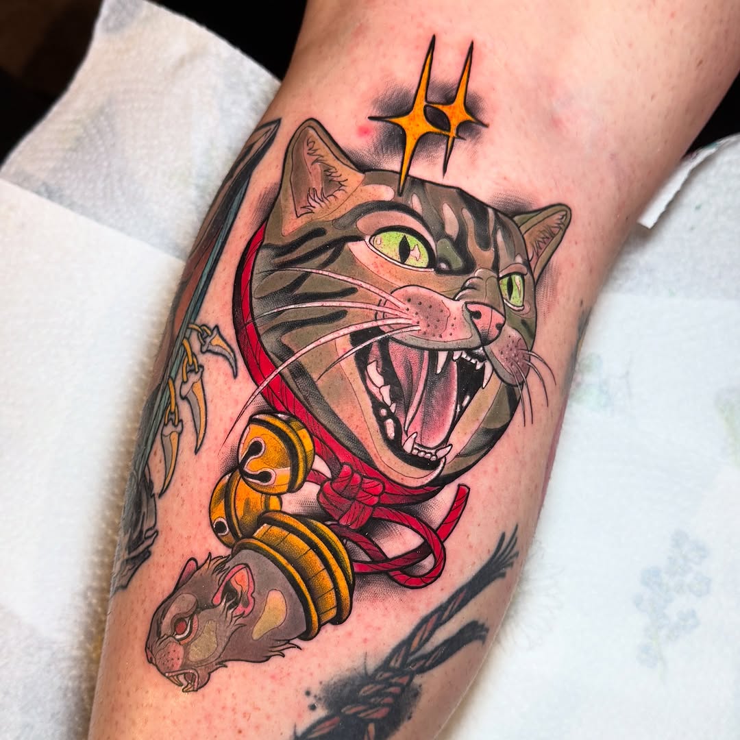 Colorful cat tattoo on calf featuring a grinning tabby with golden eyes, red collar, golden bells, and a yellow star above, surrounded by smaller decorative elements.