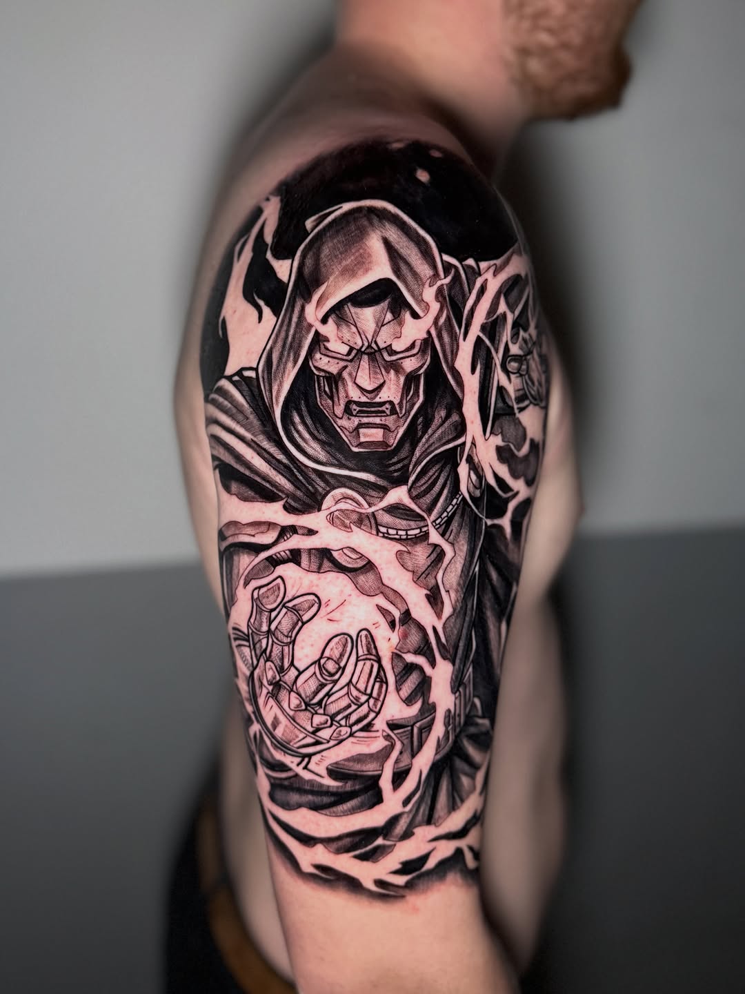 Detailed shoulder tattoo featuring a hooded skeletal figure with dramatic facial features and an outstretched skeletal hand with flowing elements in black and grey ink.