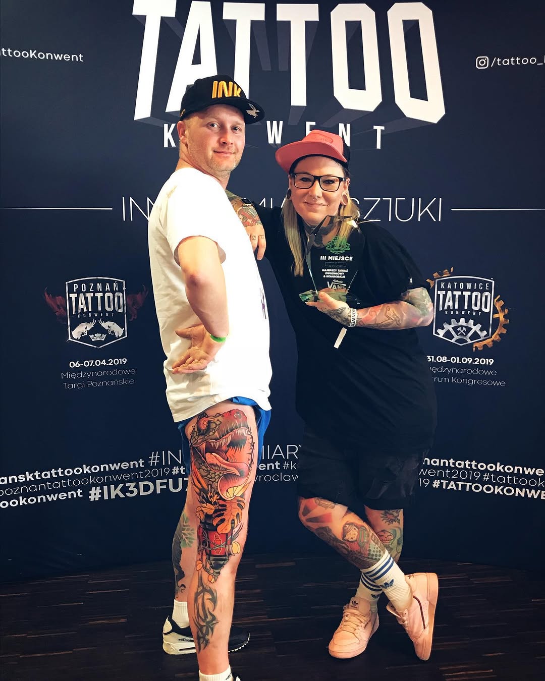 Two people pose at a tattoo convention with colorful leg and arm tattoos featuring animals, dragons, and ornamental designs in red, orange, and yellow tones.