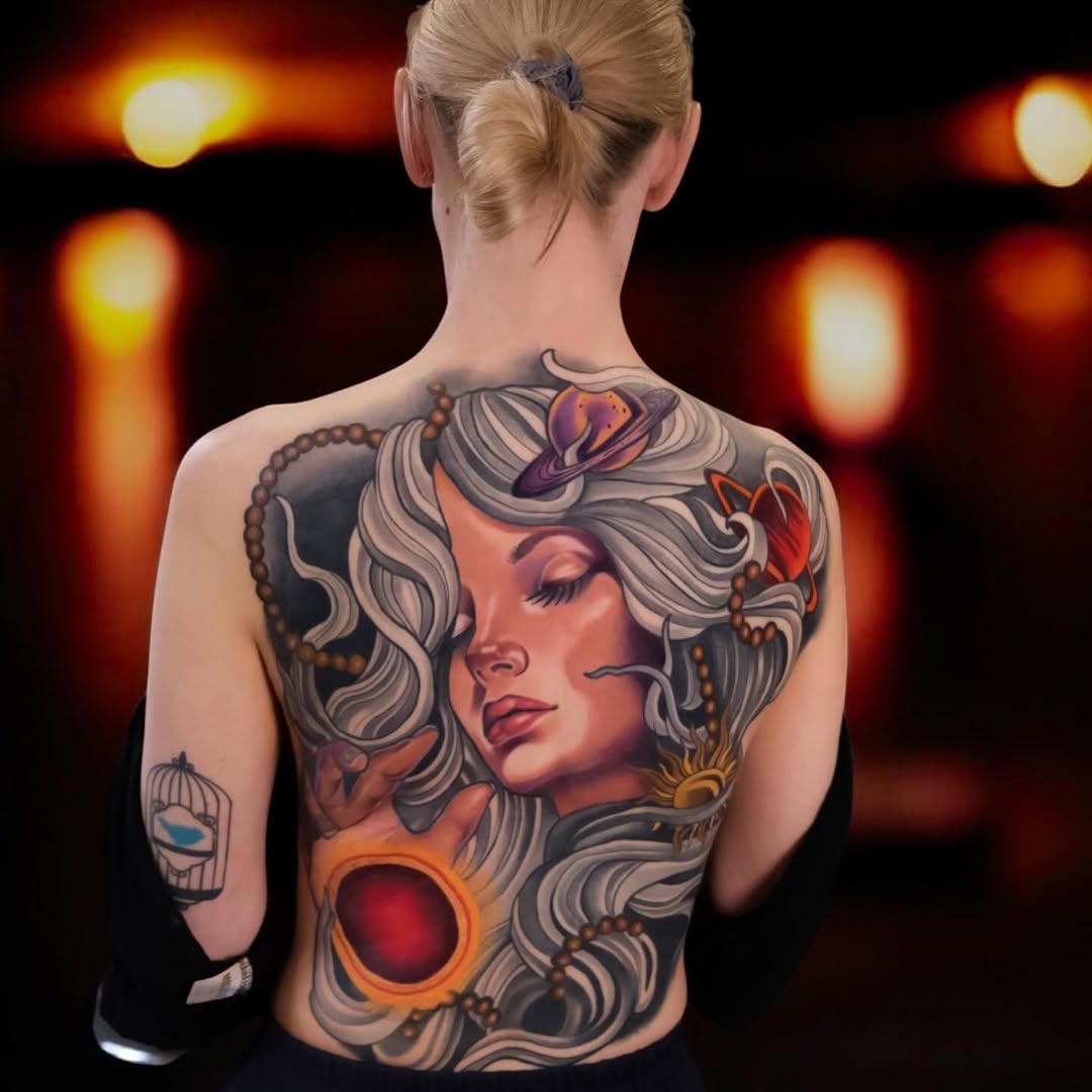 Back tattoo featuring a woman's profile with flowing hair, surrounded by decorative elements including a red orb and purple butterfly, in orange, red, and grey tones.