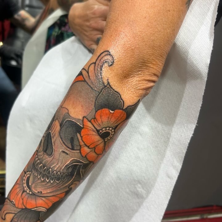 Detailed skull with orange flowers and ornamental swirls tattooed on the forearm, shown on a person wearing a white sleeve.