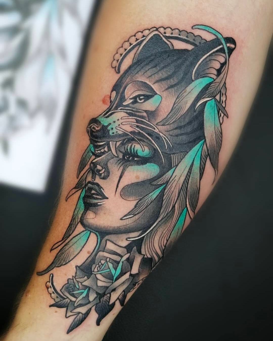 Tattoo on forearm featuring a lion's head above a woman's face with flowing hair, accented with turquoise color and floral elements at the base.