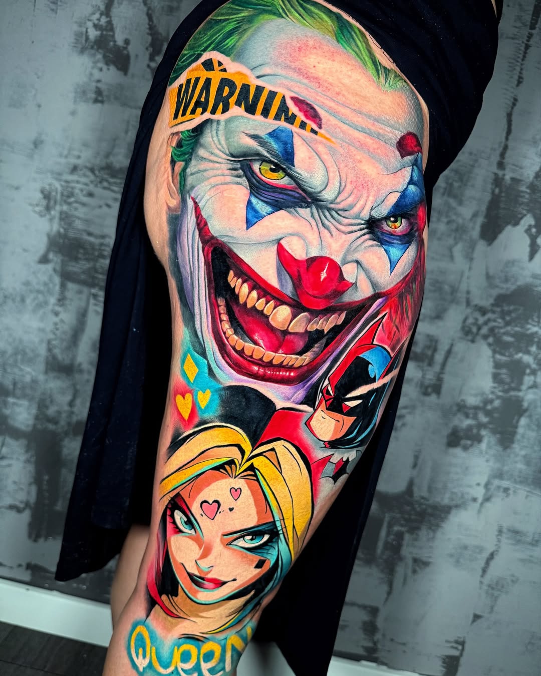 Colorful upper arm tattoo featuring a menacing clown face with a "warning" banner, a jester character, and a blonde woman with hearts, predominantly red, blue, yellow, and green.