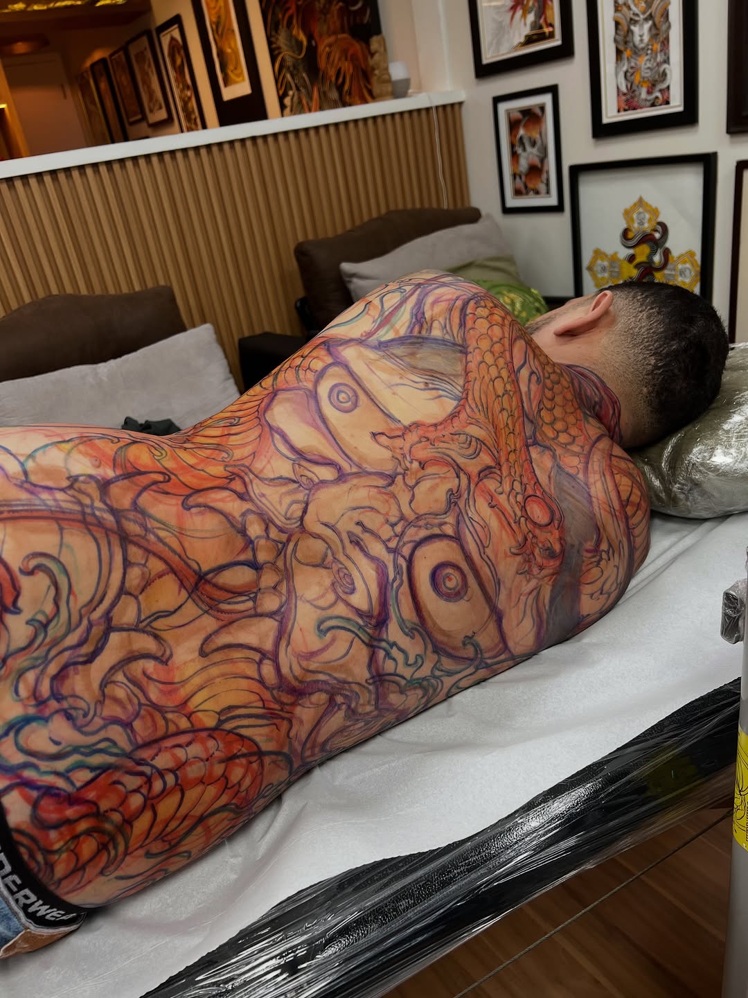 Full-back tattoo featuring intricate organic patterns in orange, purple, and red tones covering the torso and shoulders of a client lying face-down.