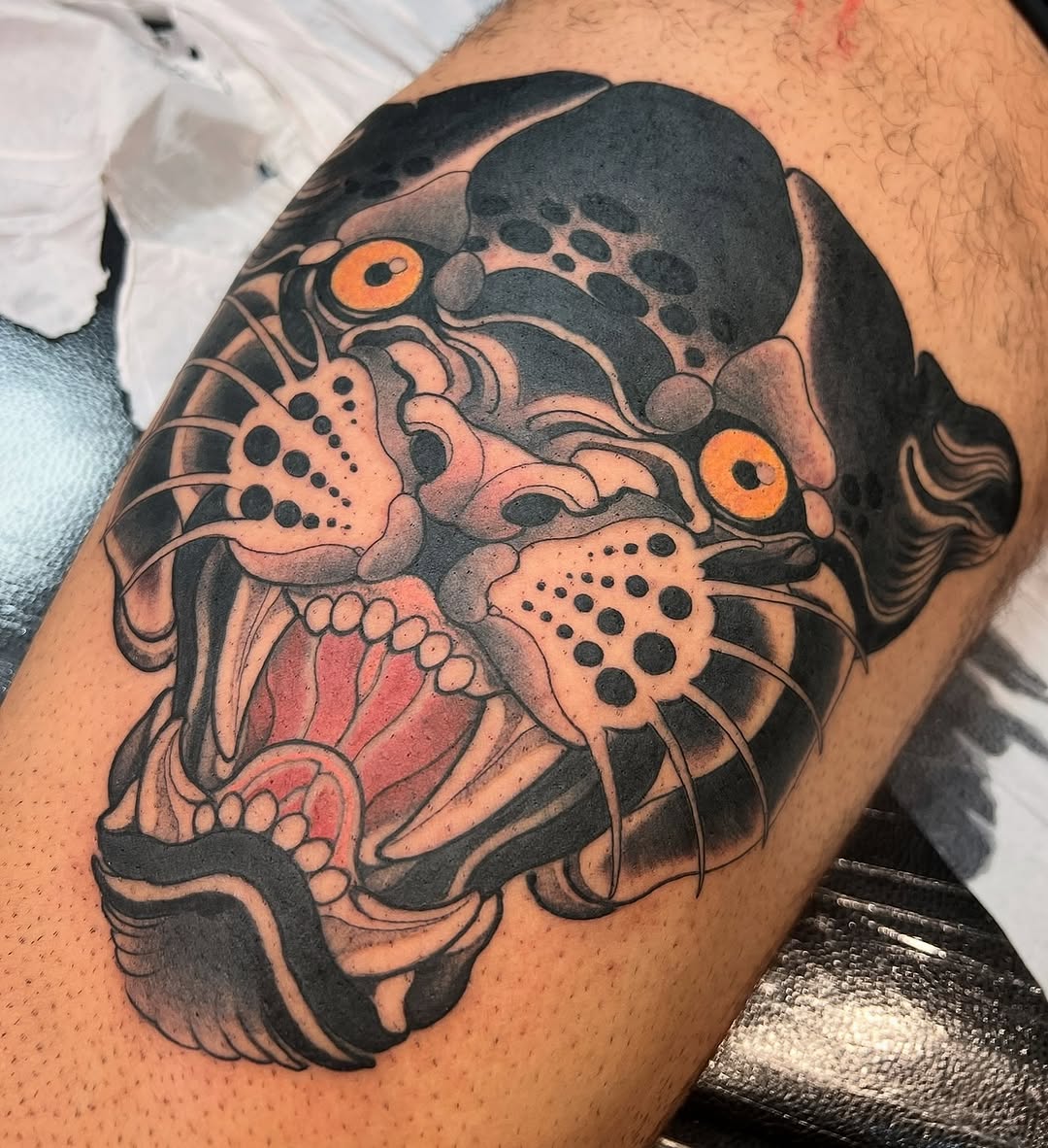 Colorful demon or oni mask tattoo with orange eyes, sharp teeth, whiskers, and ornate details on upper arm in black, orange, and flesh tones.