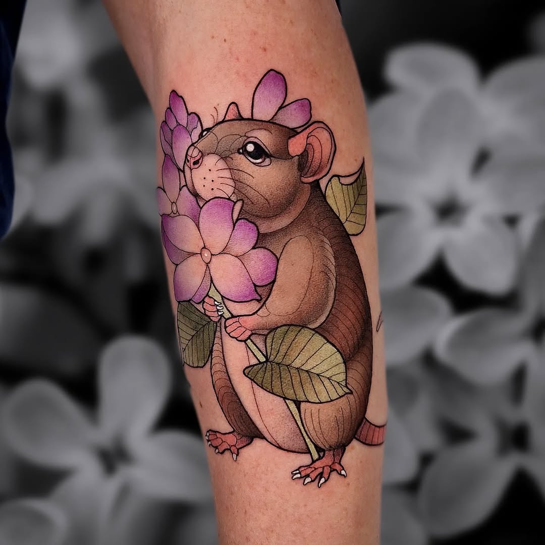 Colorful tattoo on an arm depicting a brown mouse holding purple flowers with green leaves, featuring detailed linework and shading.