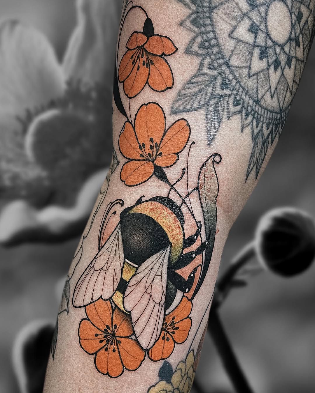 Colorful tattoo on an arm featuring orange flowers, a detailed bee with black and gold wings, green leaves, and surrounding botanical elements.