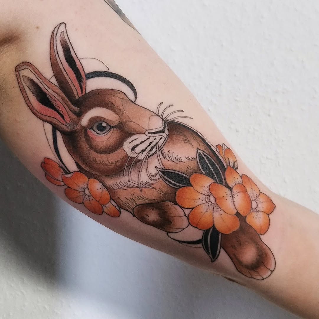 Brown rabbit with pink ears and whiskers holding orange flowers, tattooed on an upper arm with warm brown and orange coloring.