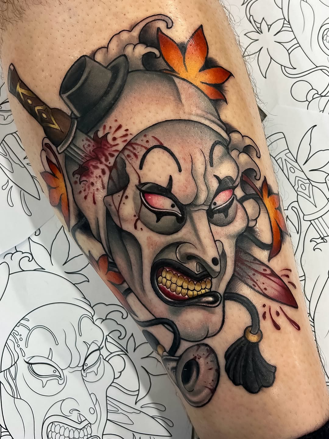 Demon mask tattoo with golden teeth, red eyes, orange flowers, and blood drips on the upper arm in color and black ink.