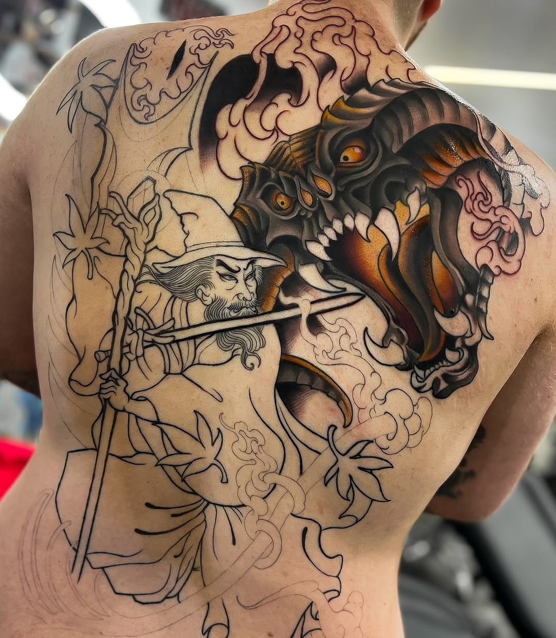 Back tattoo featuring a demon head with golden eyes and orange accents facing a bearded man's profile, surrounded by ornamental scrollwork and floral designs.