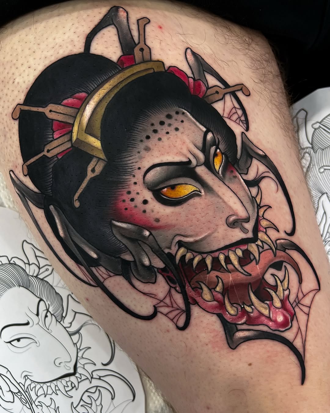 Colored shoulder tattoo of a demonic face with yellow eyes, black horns, red details, and ornate headpiece with weapons.