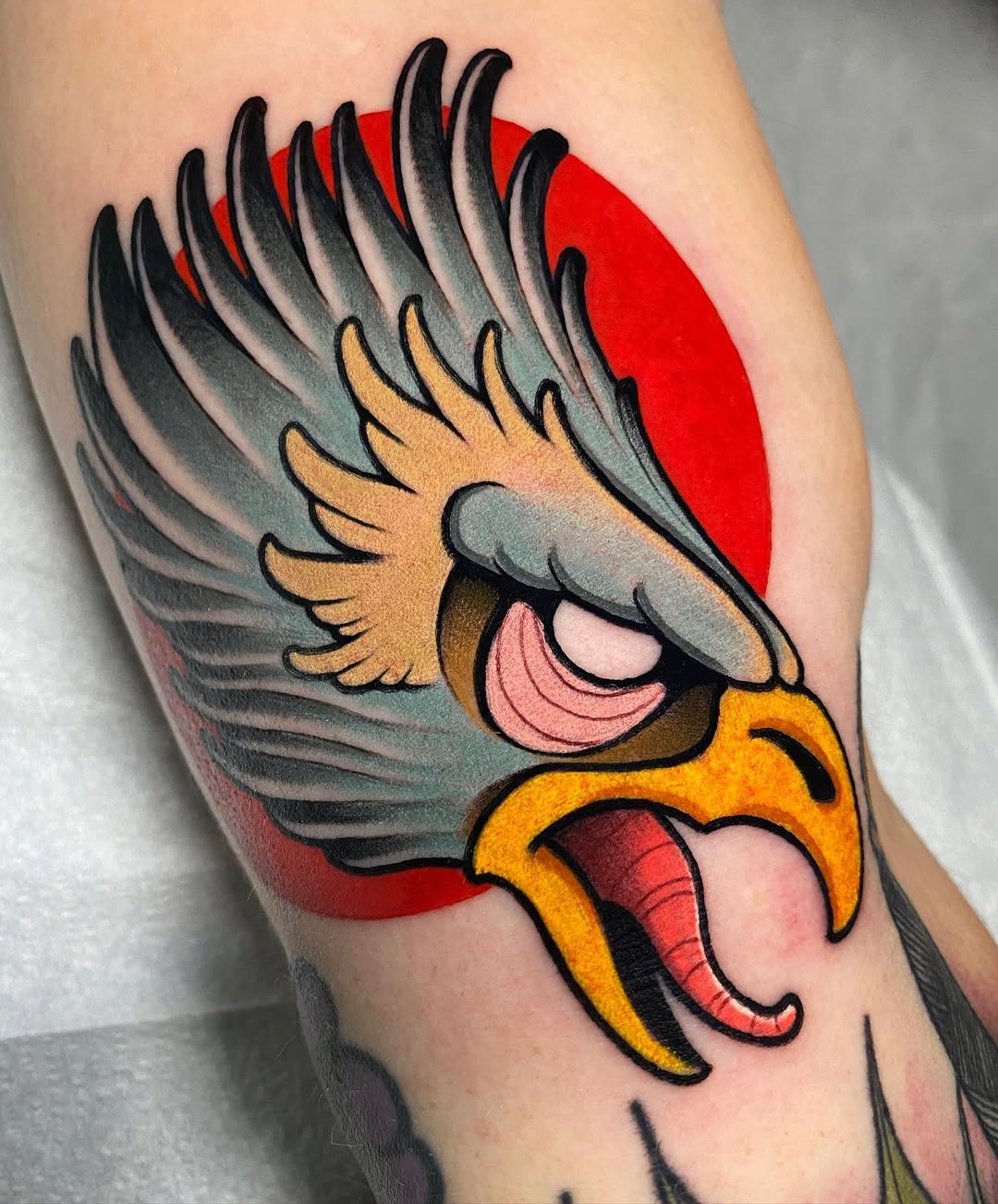 Eagle head with spread wings and open beak in yellow, black, and gray tones against a red circular background on upper arm.