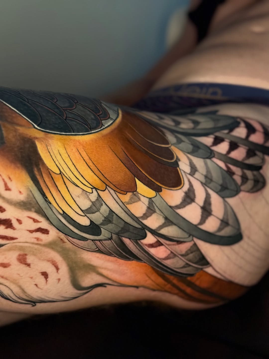 Colorful bird wing tattoo with golden-yellow and black feathers on the upper arm, featuring detailed feather line work and shading.