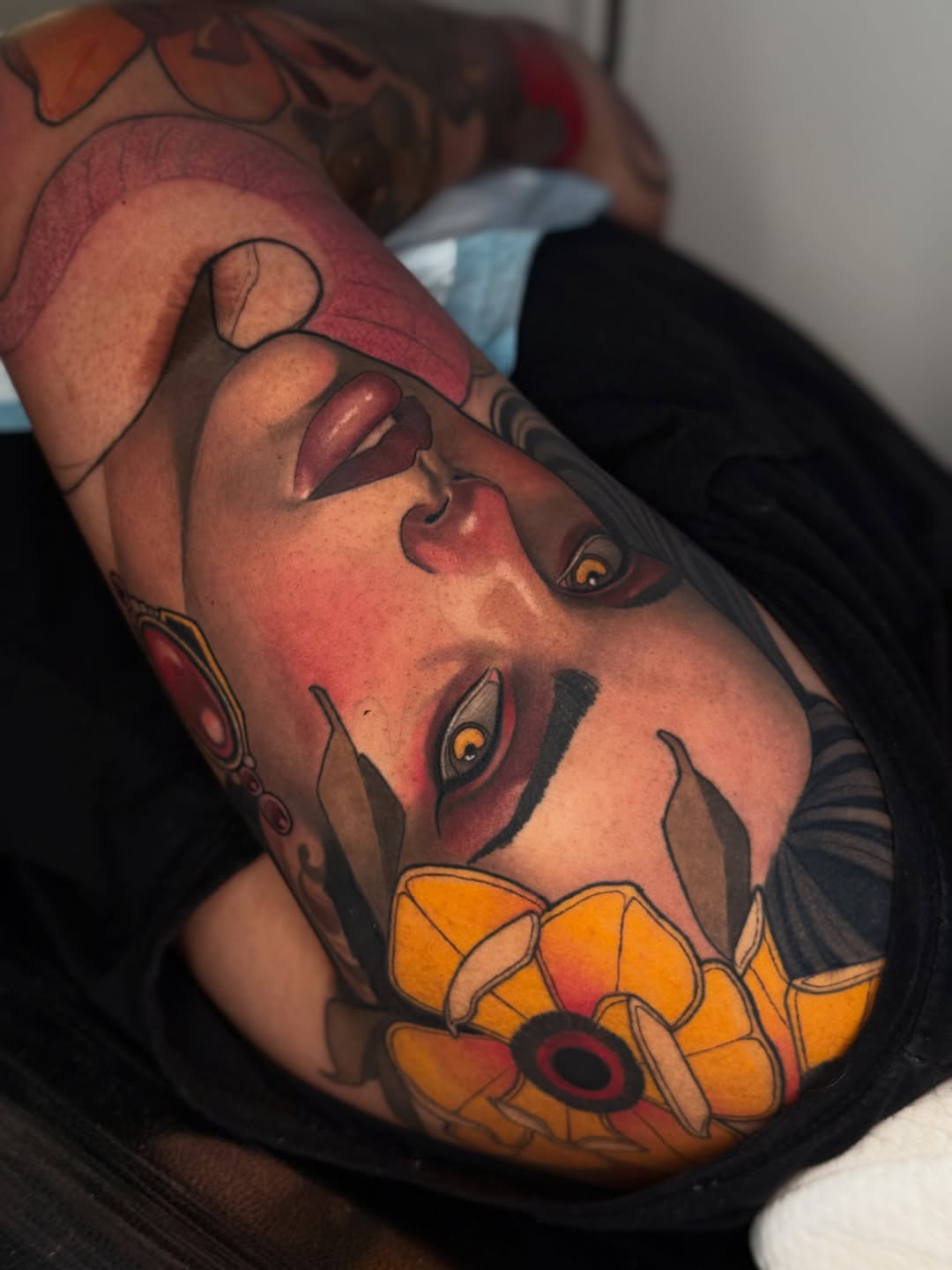 Colorful portrait tattoo on forearm featuring a face with expressive eyes, red and burgundy tones, and a yellow flower accent at the base.