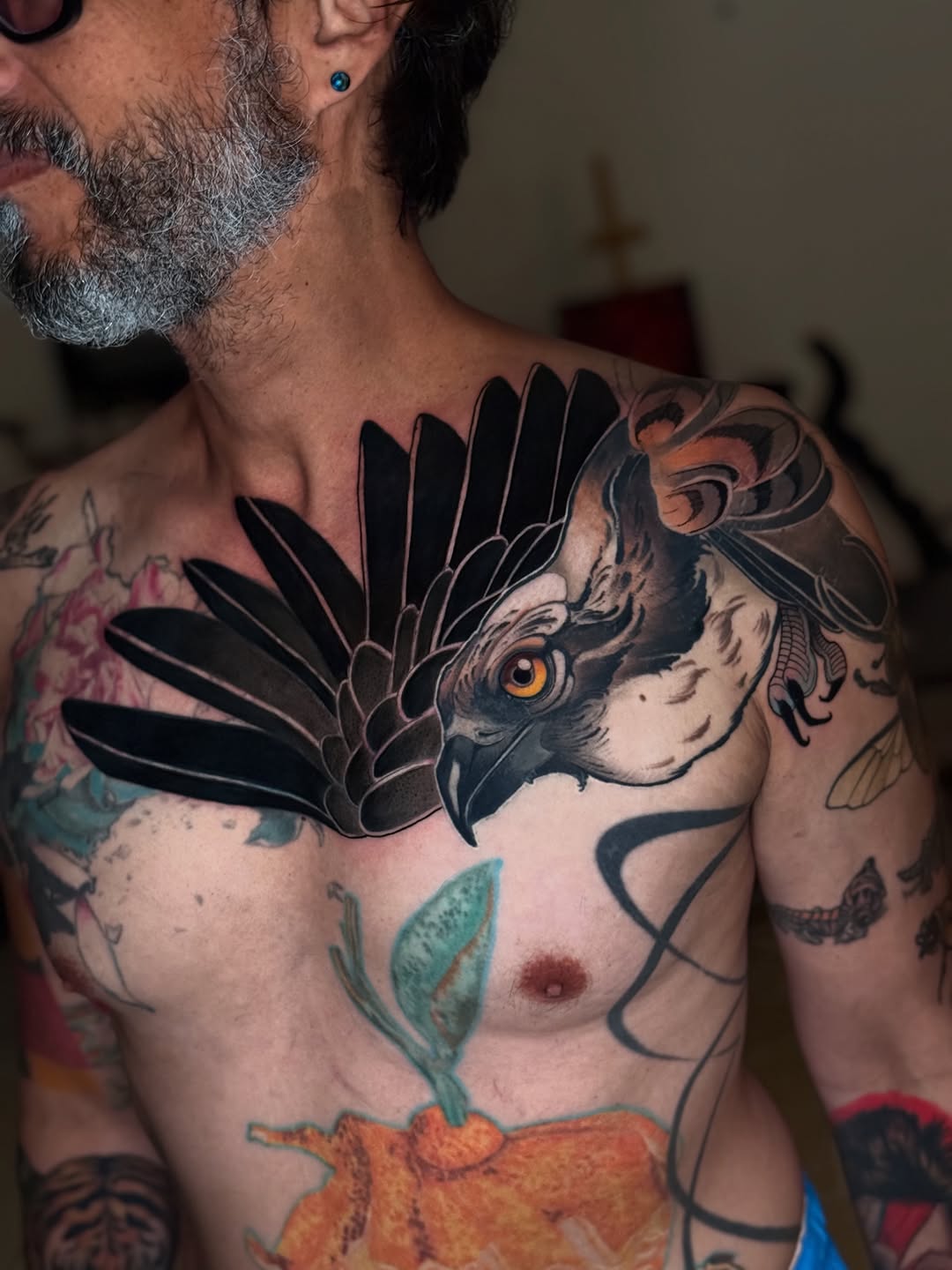 Cockatoo head tattoo with black feathers and golden eye on chest, surrounded by orange flower and green leaves.