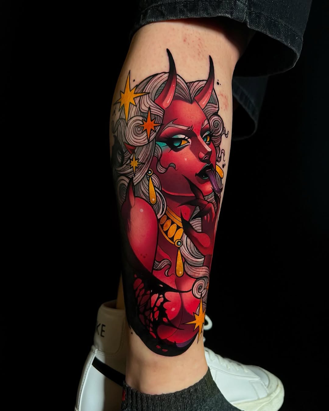 Red devil head with horns, wavy hair, teal eyes, gold stars and trident adorning the left calf muscle.
