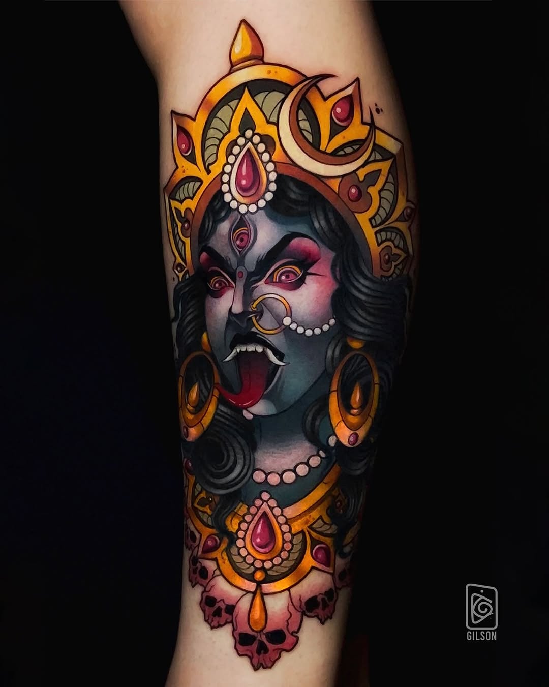 Colorful tattoo on a leg featuring a dark-faced deity with ornate gold crown, jeweled accessories, and decorative floral borders in orange, red, and cream tones.