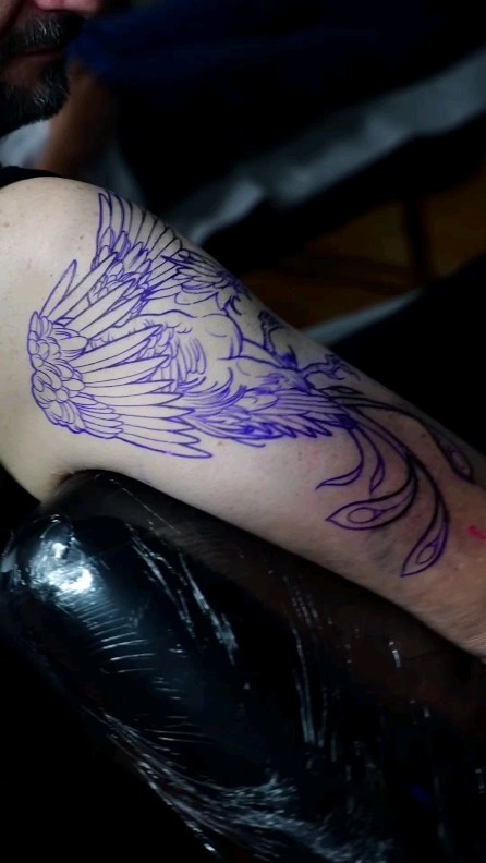 Purple-outlined winged phoenix design tattooed on upper arm and shoulder, with flowing tail extending down the forearm.
