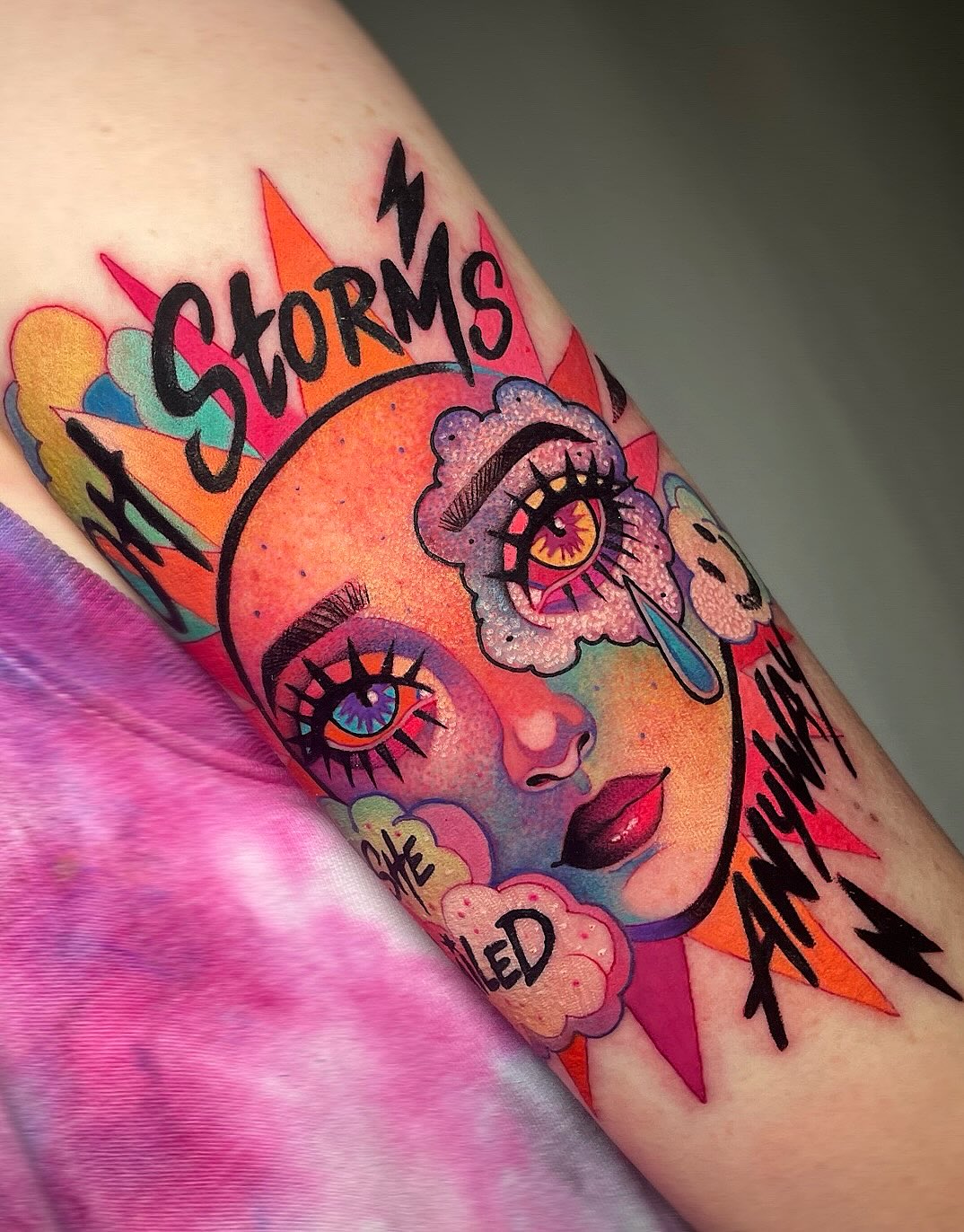 Colorful face tattoo on upper arm featuring "Storms" text, expressive eyes with long lashes, flowers, lightning bolts, and vibrant orange, pink, purple, and yellow hues.