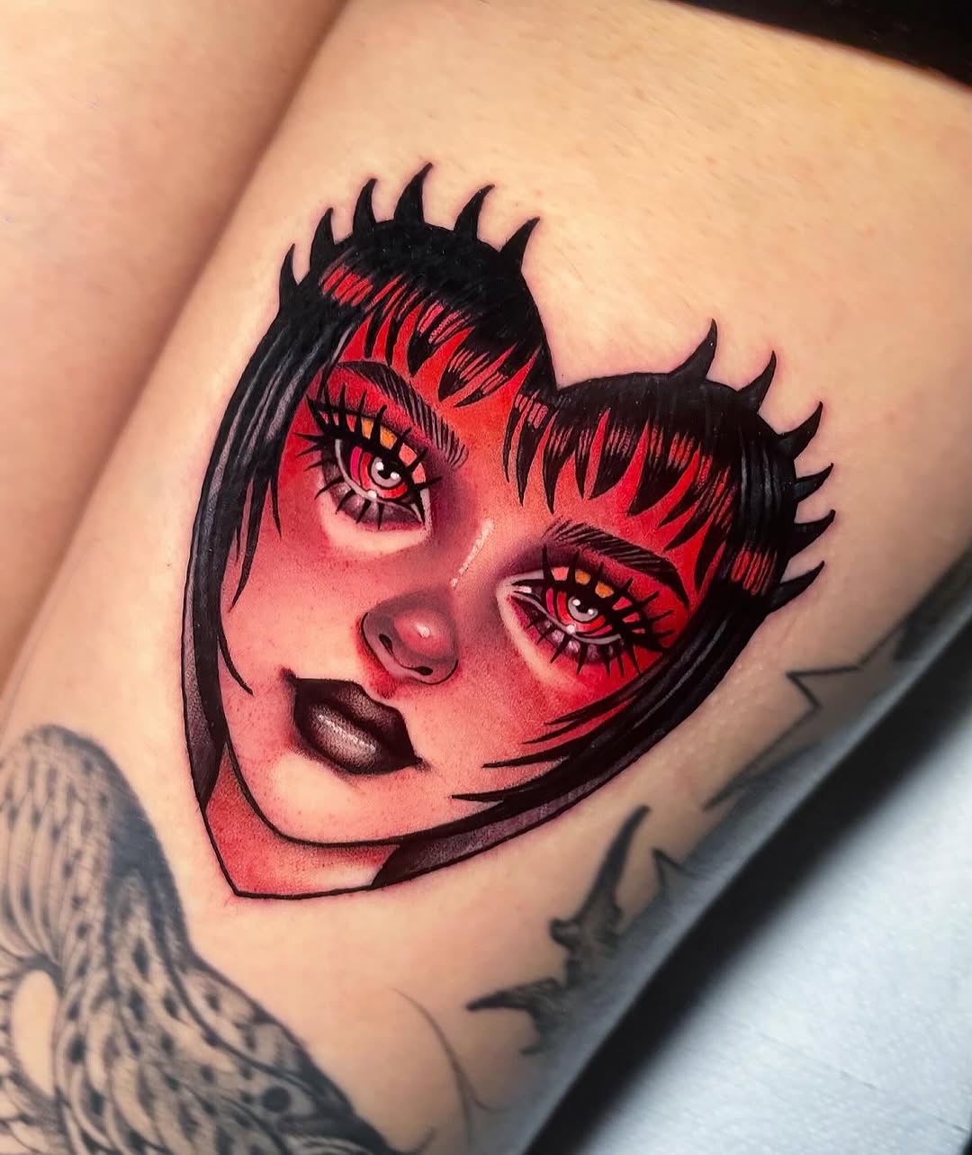 Red and black portrait tattoo of a stylized female face with pointed ears, dramatic eyes with spiked lashes, and bold lips on the upper arm.