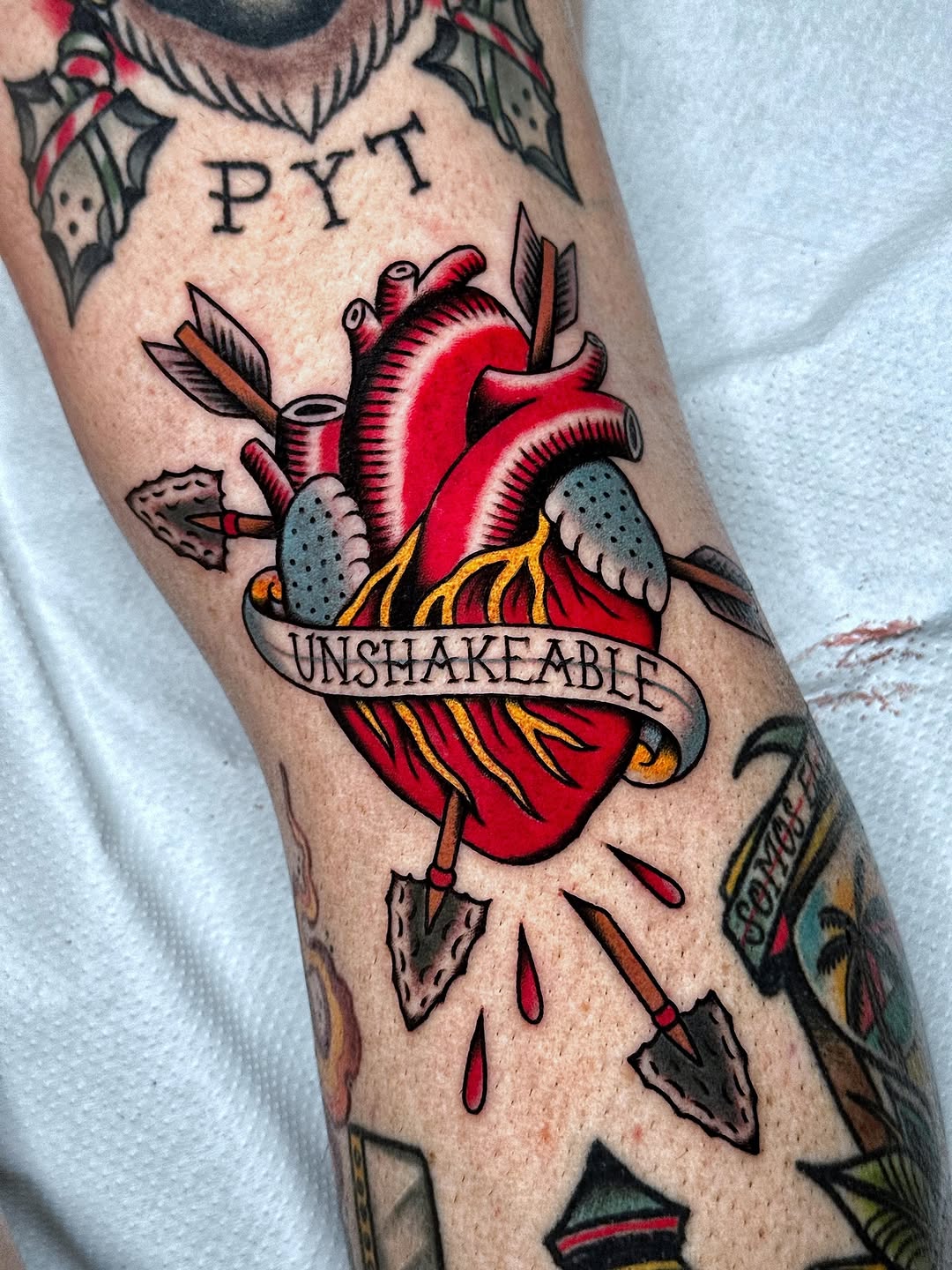 Red anatomical heart pierced by arrows with gray wings and "UNSHAKEABLE" banner on inner forearm, surrounded by leaf and drop details.