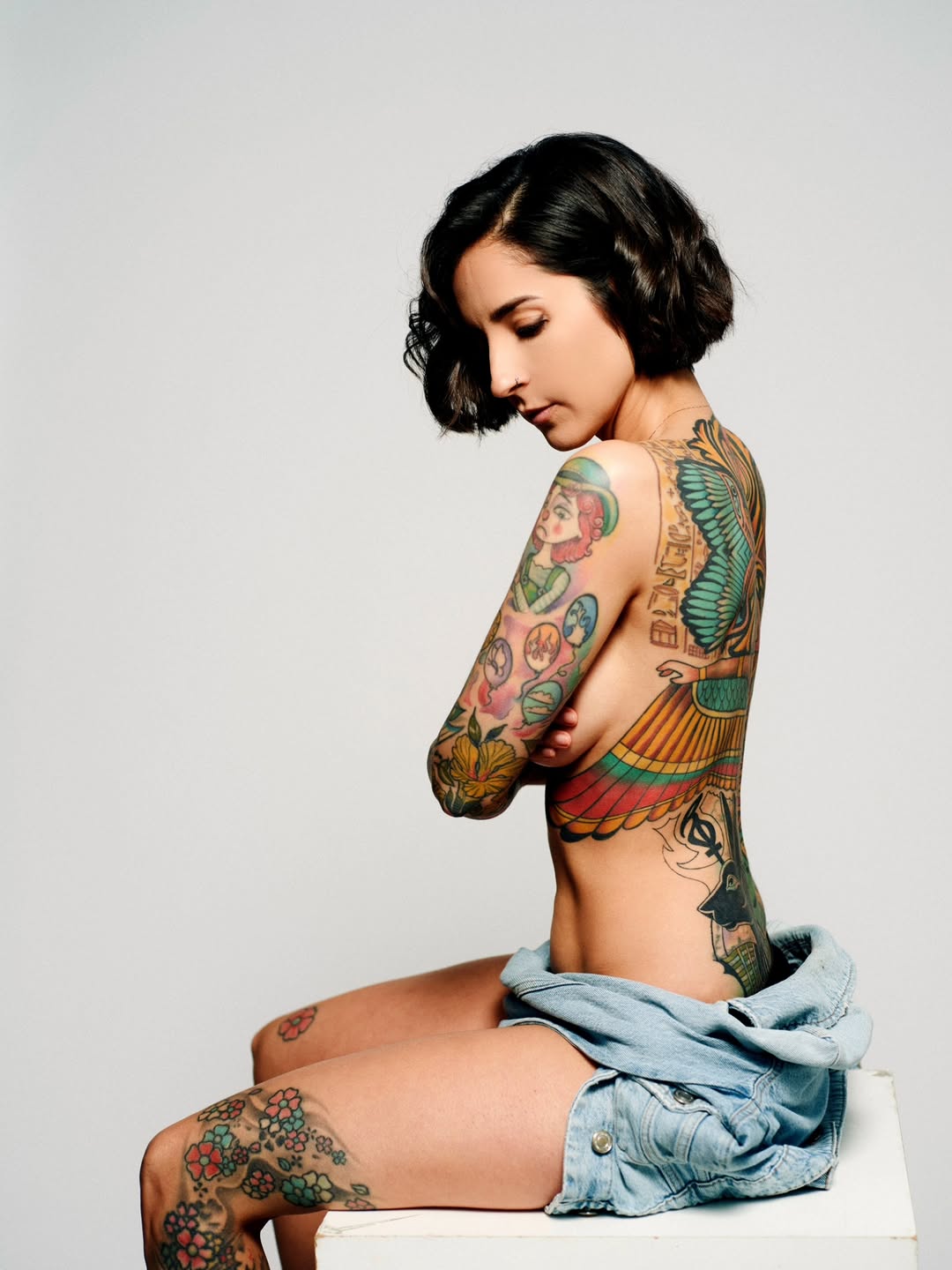Woman displaying extensive colorful body tattoos including Egyptian imagery, winged figures, and floral designs on arms, torso, and legs in portfolio photograph.