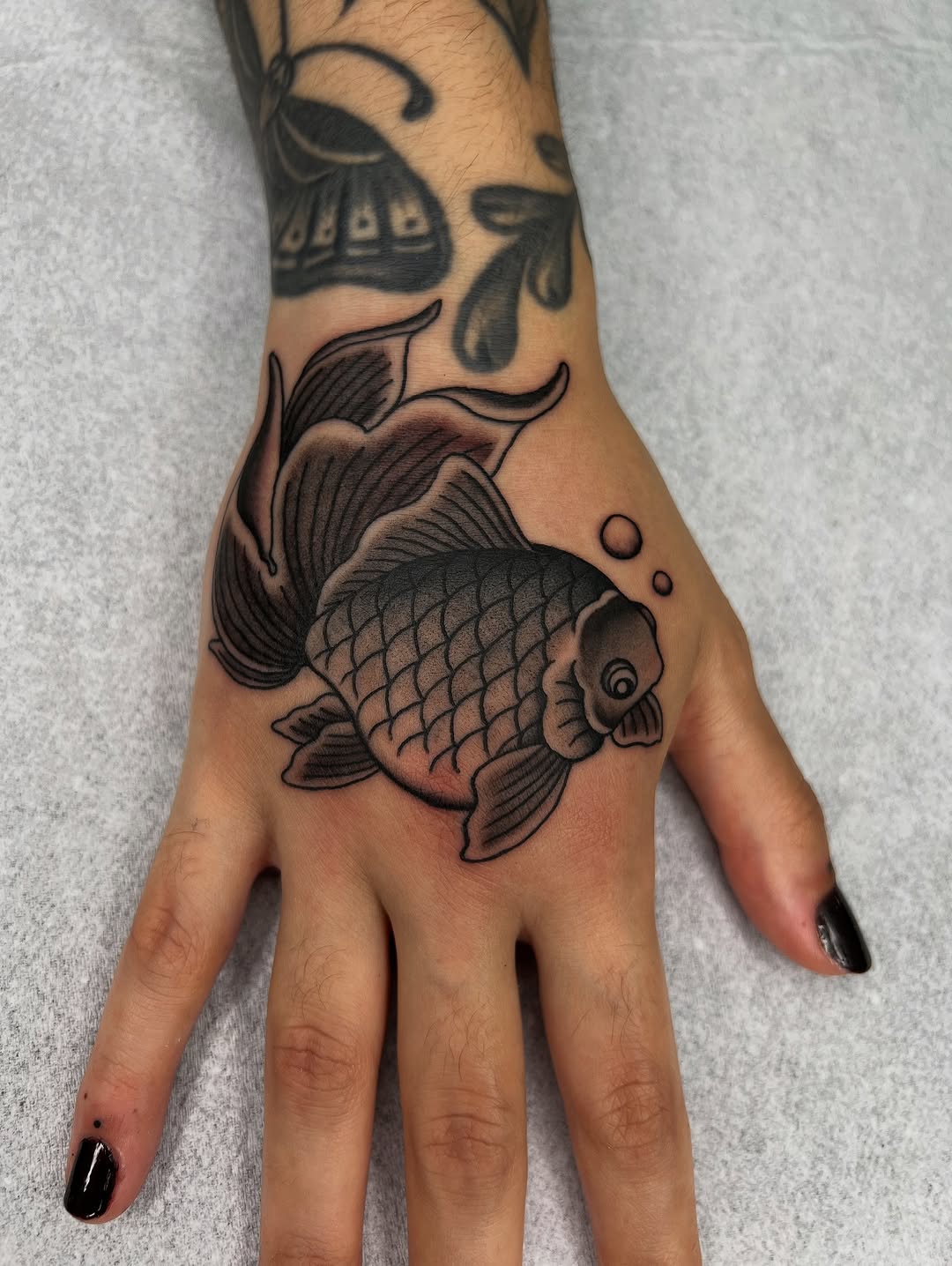 Fish tattoo on the back of a hand featuring detailed scales, flowing fins, and shading in black and brown ink with bubbles.