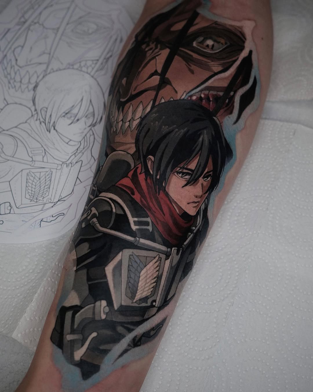 Colored tattoo on upper arm featuring two anime characters in dark clothing with detailed facial features and armor elements, displayed next to sketch designs.