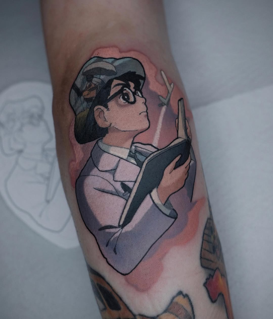 Colored tattoo on forearm depicting a person wearing a cap and holding an open book, rendered in muted pink, gray, and black tones.