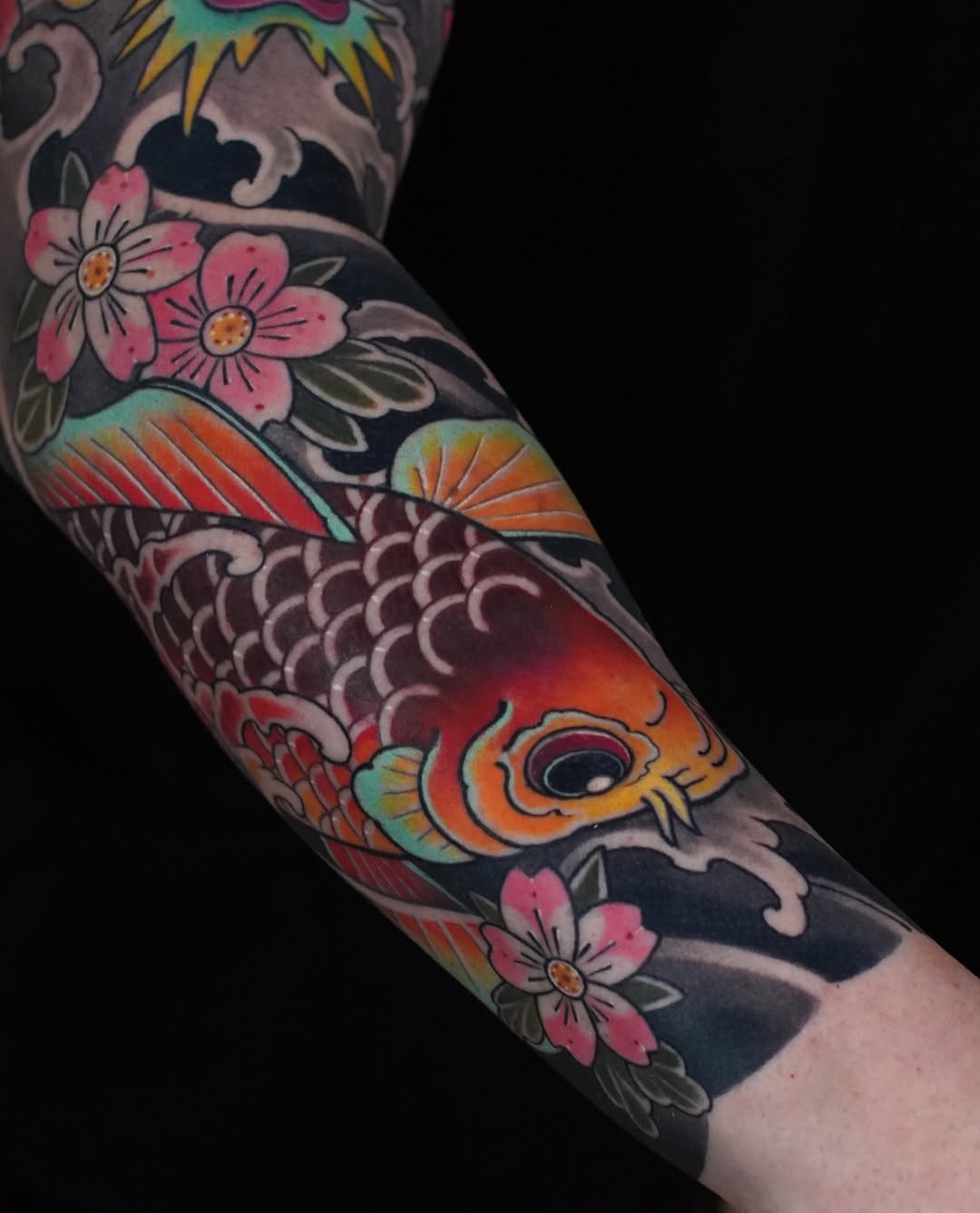 Colorful koi fish tattoo on forearm with pink flowers, orange and red scales, and decorative waves on dark skin background.